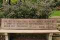 A wood park bench engraved with an inspirational quote in front of a stone retaining wall and shrubs in Janesville, Wisconsin