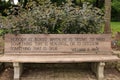 A wood park bench engraved with an inspirational quote in front of a stone retaining wall, shrubs and trees