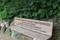 A wood park bench engraved with a quote from George Santayana in front of a large shrub and a rock retaining wall