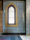 Wooden arched window framed by golden floral pattern ornaments over ceramic tiles wall with floral blue patterns