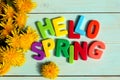 On a wooden background there is an inscription in multi-colored letters hello spring surrounded by dandelion flowers