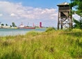 Wooden bird watch tower and port with commercial ships and cranes. Grass and sky with clouds