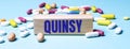 A wooden block with the word QUINSY stands on a blue background among multi-colored pills. Medical concept
