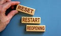 Wooden blocks form the words `reset, restart, reopening` on blue background. Male hand
