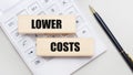 Wooden blocks with the LOWER COSTS Iie on a light background on a white calculator. Nearby is a black handle. Business concept