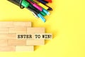 Wooden blocks stacked next to pens and pencils on a yellow background. ENTER TO WIN text on a wooden block.