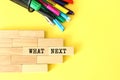 Wooden blocks stacked next to pens and pencils on a yellow background. WHAT NEXT text on a wooden block.