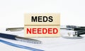 Wooden blocks with text Meds Needed on a clipboard, stethoscope and pen