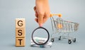 Wooden blocks with the word GST Goods & Services Tax, money and a supermarket trolley. Tax, which is imposed on the sale of