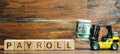 Wooden blocks with the word Payroll, money and a forklift. Payroll is the sum total of all compensation a business must pay to its