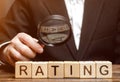 Wooden blocks with the word Rating and a magnifying glass in the hands of a businessman. The concept of studying the rating of the