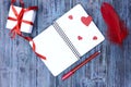 On a wooden blue table lies an open empty notebook, red pen, red bird feather and several red hearts and a white box with a gift. Royalty Free Stock Photo