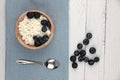 Wooden bowl of farmer cheese with a spoon on blue napkin and a pile of blueberry on white wooden background flat lay. Healthy