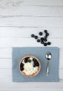 Wooden bowl of farmer cheese with a spoon on blue napkin and a pile of blueberry on white wooden background flat lay. Healthy