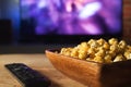 A wooden bowl of popcorn and remote control in the background the TV works. Evening cozy watching a movie or TV series at home Royalty Free Stock Photo
