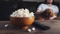 Wooden bowl with salted popcorn and TV remote on wooden table. In the background, a man with a red dog watching TV on