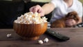 Wooden bowl with salted popcorn and TV remote on wooden table. In the background, a man with a red dog watching TV on the couch Royalty Free Stock Photo