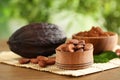 Wooden bowls of cocoa beans and powder near pod on table against blurred green Royalty Free Stock Photo