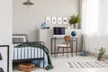 Wooden box under single metal bed with blue sheets and green blanket in trendy kid`s room with workspace Royalty Free Stock Photo