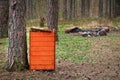 Wooden brigh orange trash bin in forest resting spot between tree trunks on green grass in spring