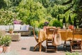 Wooden chairs at Dining table covered with orange tablecloth standing on wooden terrace in garden