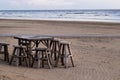 Wooden chairs and table stand on a wide sandy seashore with a coastline in perspective