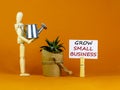 Wooden clothespin with white sheet of paper. Text `grow small business`. Wooden manequin, house plant from, miniature watering c