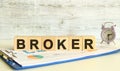 Wooden cubes lie on a folder with financial charts on a gray background. The cubes make up the word BROKER