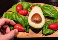 A wooden cutting board on a black background with an avocado in the center. All around, intense green basil leaves and bright red Royalty Free Stock Photo