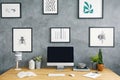 Wooden desk with empty mockup monitor, keyboard, lamp, fresh plants and notebooks in home office room interior with grey raw wall