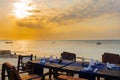 Wooden dinning table woth chairs on the beach on Zanzibar, with orange sunset, silhouettes of boats in the background. Royalty Free Stock Photo