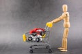 A wooden doll carries a red car model in metal shopping cart. Man buy his new car in dealership. Concept of buying new cars, Royalty Free Stock Photo