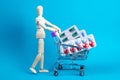 Wooden doll and metal shopping cart with pills on blue background. Various capsules, tablets and medicine on blue pastel trendy