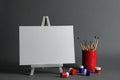 Wooden easel with blank canvas board and painting tools for children on dark background.