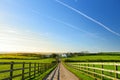 Fence casting shadows on a road leading to small house between scenic Cornish fields under blue sky, Cornwall, England