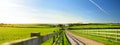 Fence casting shadows on a road leading to small house between scenic Cornish fields under blue sky, Cornwall, England
