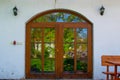 Wooden front door of a rural white house with glass reflecting the green garden and two lamp lights on both sides