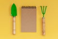 Wooden handled garden hand trowel and hand fork and empty noteboook on yellow background