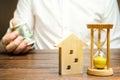 Wooden house and clock. Businessman holds money. Payment of deposit or advance payment for renting a home or apartment. Long-term Royalty Free Stock Photo