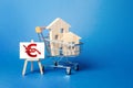 Wooden houses in a shopping cart and an easel with a red euro arrow down chart. Low sales. Value cost decrease. Bad attractiveness Royalty Free Stock Photo