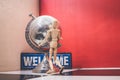 Wooden Human Manikin posing with an ornamental silver world globe and a blue welcome sign