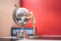 Wooden Human Manikin posing with an ornamental silver world globe and a blue welcome sign