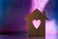 Wooden icon of house with hole in the form of heart on pink and purple dark galactic background with light bokeh
