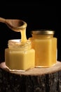 On a wooden log there are two glass jars with honey on a dark background. take honey out of the jar with a wooden spoon