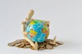 Wooden mannequin sitting with earth globe on pile of coins - Concept of planet earth protection, globalization and economy