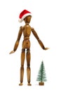 Wooden mannequin white background with Santa Claus hat his head and nearby snow covered Christmas tree.