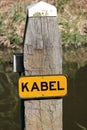Wooden mooring post in the water with yellow sign to warn that there is a cable kabel in dutch on the bottom of the water. Royalty Free Stock Photo