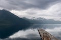 A wooden pier juts into the reflective fjord, drawing the eye towards the overcast Norwegian mountainscape