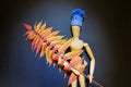 Wooden puppet doll holds a twig with autumn yellow and orange leaves, wears a blue knitted winter hat