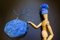 Wooden puppet wears a blue knitted winter hat and points aside with its hand at ball of yarn and crochet hook Royalty Free Stock Photo